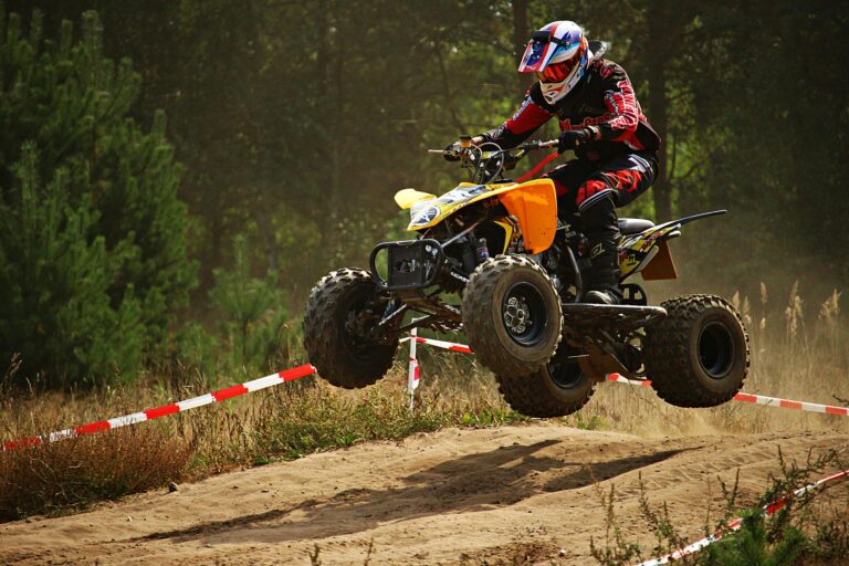 Motorsport rider performs a high-speed ATV jump on a dusty outdoor track during a race.